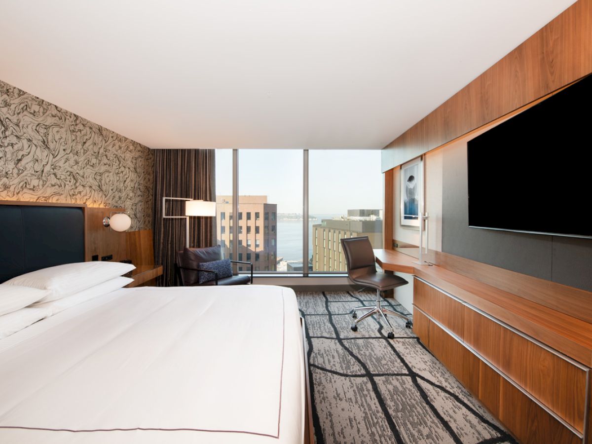 A modern hotel room featuring a large bed, flat-screen TV, desk with chair, patterned wall, and floor-to-ceiling window with a city view.