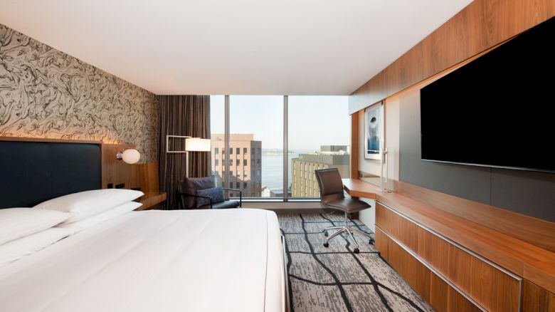A modern hotel room featuring a large bed, flat-screen TV, desk with chair, patterned wall, and floor-to-ceiling window with a city view.