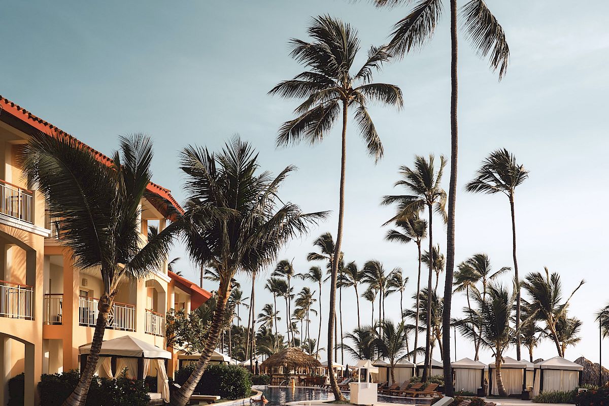 The image shows a serene pool area lined with lounge chairs, palm trees, and a resort building under a clear sky.