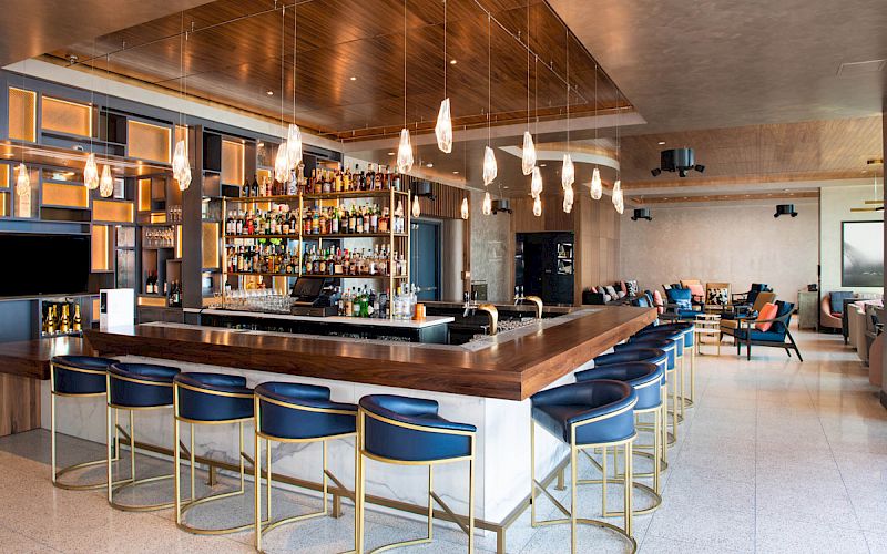 The image shows a modern bar with blue bar stools, wooden counter, a variety of liquors on shelves, and stylish pendant lights hanging from the ceiling.