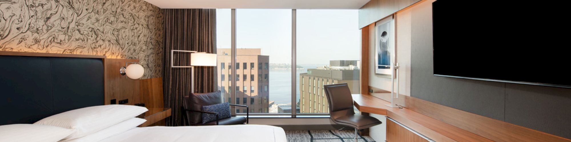 A modern hotel room features a large bed, a wall-mounted TV, a desk with a chair, and a window offering a cityscape view with buildings beyond.